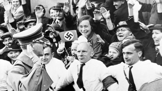 An Associated Press photograph shows Adolf Hitler, German Reichsfuehrer, acknowledging the ecstatic cheers of Sudeten Germans, as he entered Asch, on the heels of German armies which took over the ceded Czechoslovakian territory, on Oct. 3, 1938. The AP caption notes: 'Party members grip each other's belt, straining to hold the enthusiastic crowds in check.'